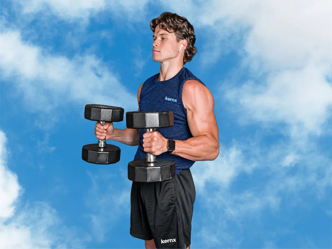 Man in a KERNX tank top lifting dumbbells against a blue sky